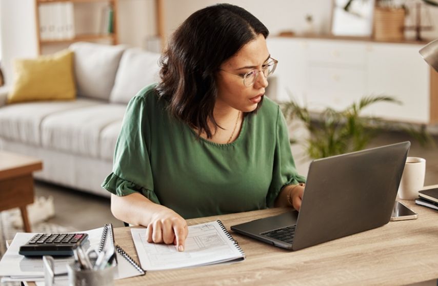 A woman works on a laptop while referencing a notebook